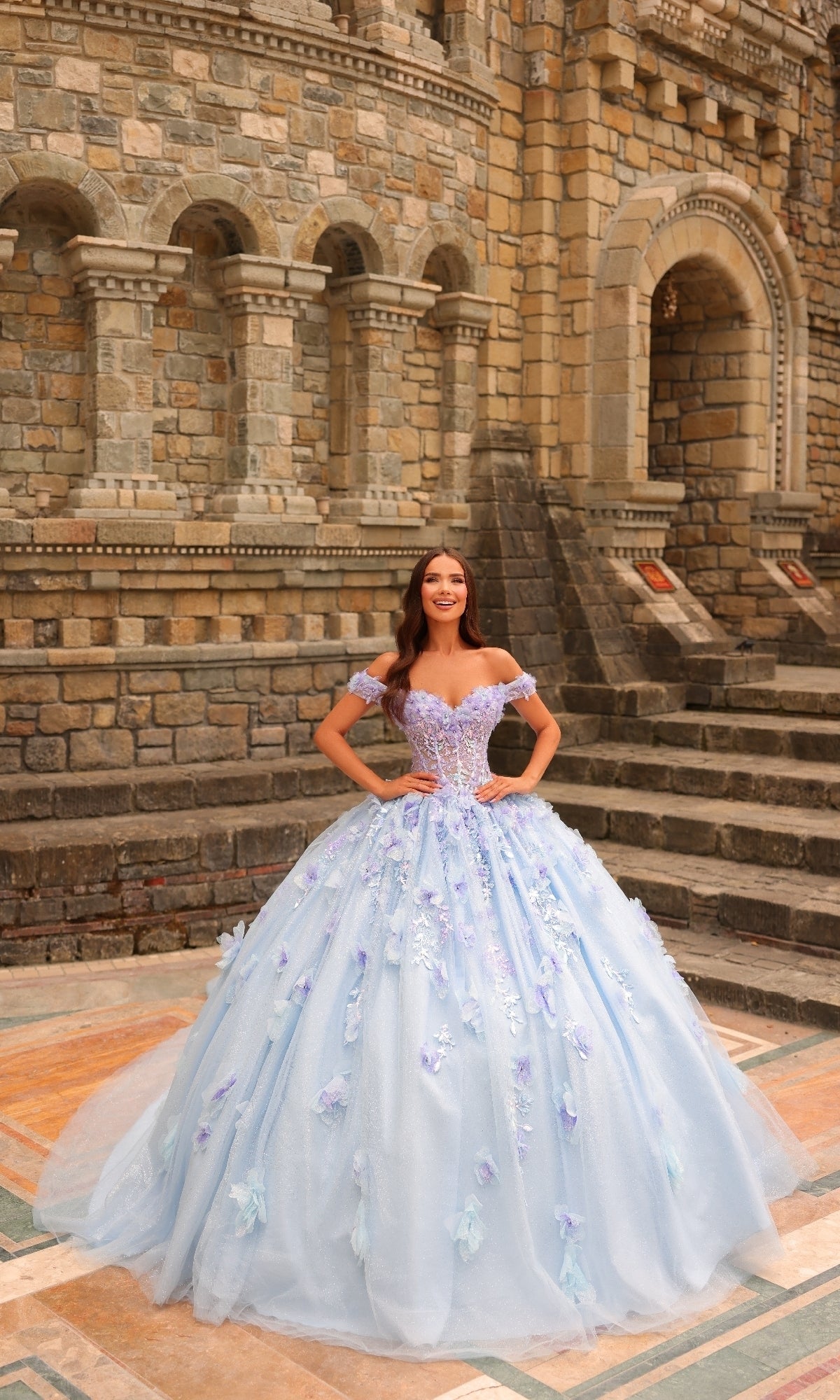 A woman smiles in front of a stone building, wearing the Amarra 54231: a light blue, off-the-shoulder 3D floral quinceañera dress.